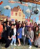 a group of cheerful, diverse individuals gathered for a photo in front of a whimsical mural depicting a town scene