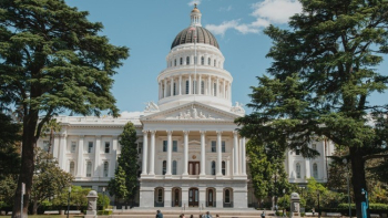 White domed capitol building framed by tall trees, with columns, steps, and people walking in front under a clear blue sky.