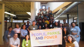 Large group of smiling people gathered on a staircase in a bright loft space, holding a "People, Parks, and Power" banner.
