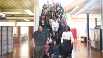 Large, diverse group of people standing and sitting on a wooden staircase in a bright office, smiling toward the camera.