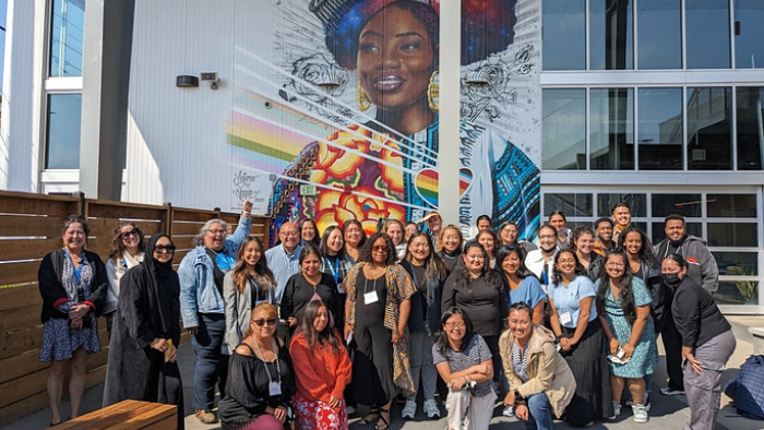 Group of diverse adults gathered outdoors, smiling in front of a large mural of a Black woman at a community center.